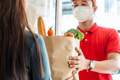 A masked delivery worker handing a bag of groceries to a woman in her doorway.