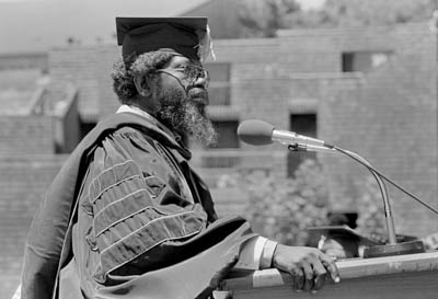 J. Herman Blake speaks at a podium wearing a cap and gown in front of Oakes College