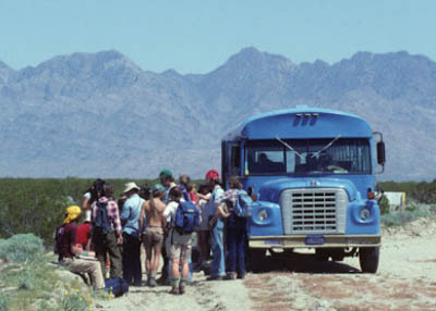 Students outside a blue bus in the desert with mountains in the background