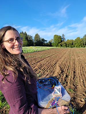 Colleen Josephson holds a microbial fuel cell in front of a plowed row of dirt.