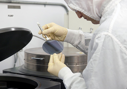 A person with gloved hands and clean suit uses a tool to hold a silicon hardware chip.