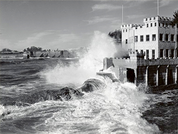 A wave crashes against a castle structure in this black and white photo.