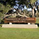 A sign shows the name University of California Santa Cruz in front of a tree on campus