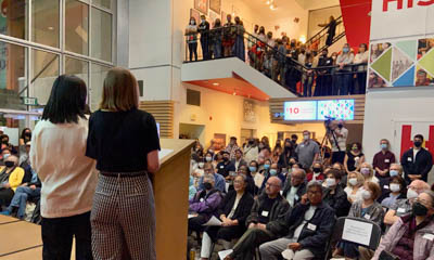 The view from behind the podium as two speakers address a large crowd in an atrium 