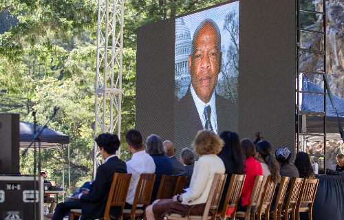 Speakers sit onstage beneath a large screen showing an image of the late Rep. John R. Lewi