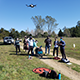 Students stand around a drone hovering in the air in a field.