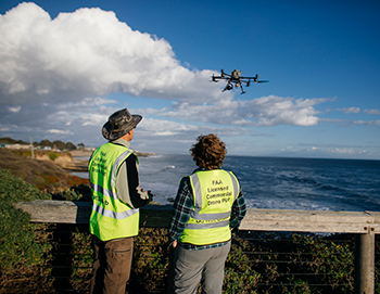 Two people in safety vests operate a drone by the coast.
