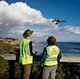 Two people in safety vests operate a drone by the coast.