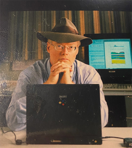 David Draper looks at a camera, sitting behind a computer and wearing a gray fedora.