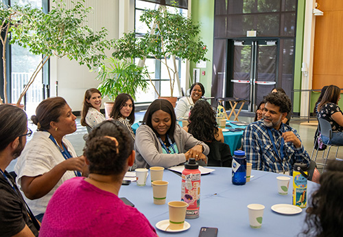 Five people chat around a table while others look on.