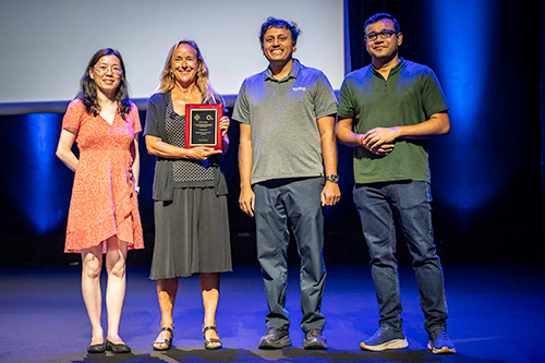 Getoor smiles on a stage holding an award plaque with three others