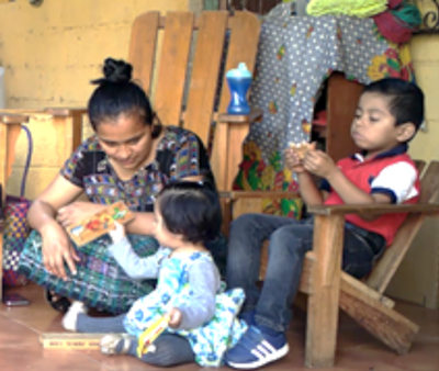 A mother and toddler play with a toy while older child in a chair plays independently