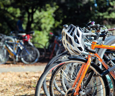Close-up of an orange bicycle parked in a bike rack with a silver helmet hanging from the handlebars.