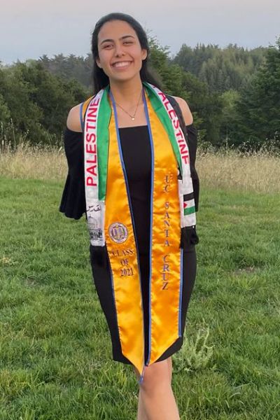 Nabeela Ariqat smiles at the camera. She wears a black dress and a yellow UC Santa Cruz Class of 2021 stole and a Palestinian flag stole. Trees and grass fields are visible in the background.