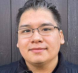 Headshot of Steven. He has short dark hair and glasses and is standing in front of a dark wooden wall. He is wearing a black jacket and looking at the camera with a neutral expression.
