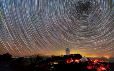 A long-exposure image captures circular star trails above telescope domes illuminated in red light on a dark mountain. A golden glow on the horizon marks distant city lights.