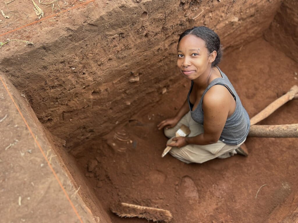 Venerable kneels inside a rectangular hole in red dirt with a brush she has used to uncover bones