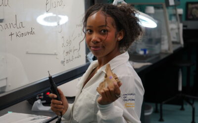 Elyse Venerable holds a saw blade and horse jawbone with teeth in a lab while wearing a white lab coat