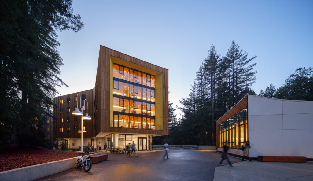 modern, multi-story building at dusk on the UC Santa Cruz campus, with warm interior lighting glowing through large glass windows. Students walk and bike through the plaza surrounded by tall redwood trees, with a second, lower building on the right also illuminated inside.