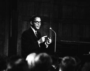 Black and white photo of Tom Lehrer sitting next to a piano on stage, speaking to the audience