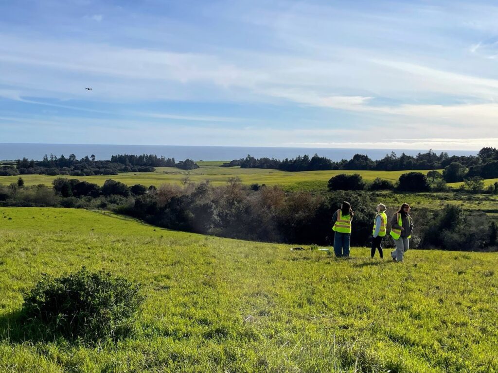 From far away, three figures fly a drone on the UC Santa Cruz campus with a view of the Monterey Bay in the background.