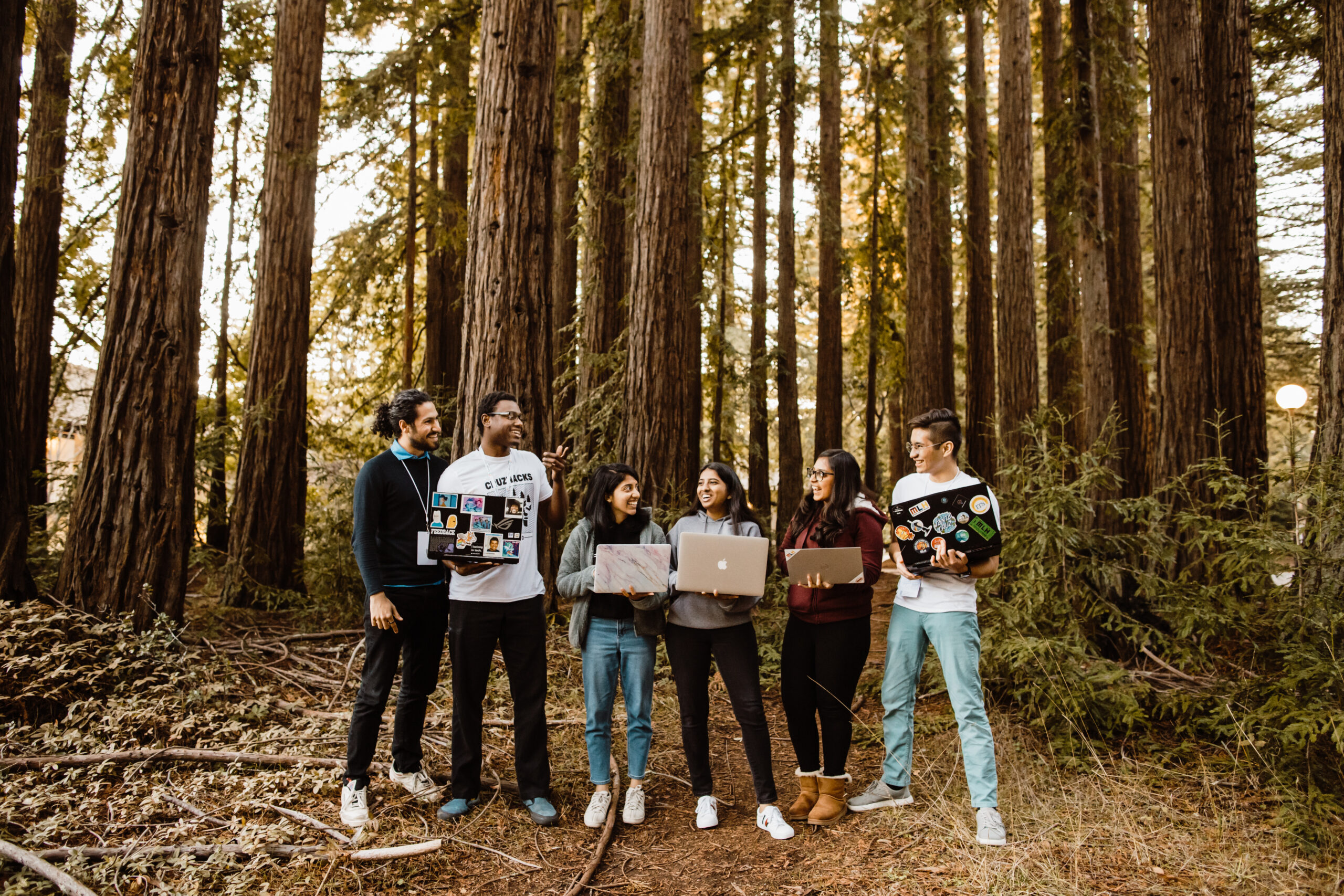 Students stand in front of redwood trees holding computers