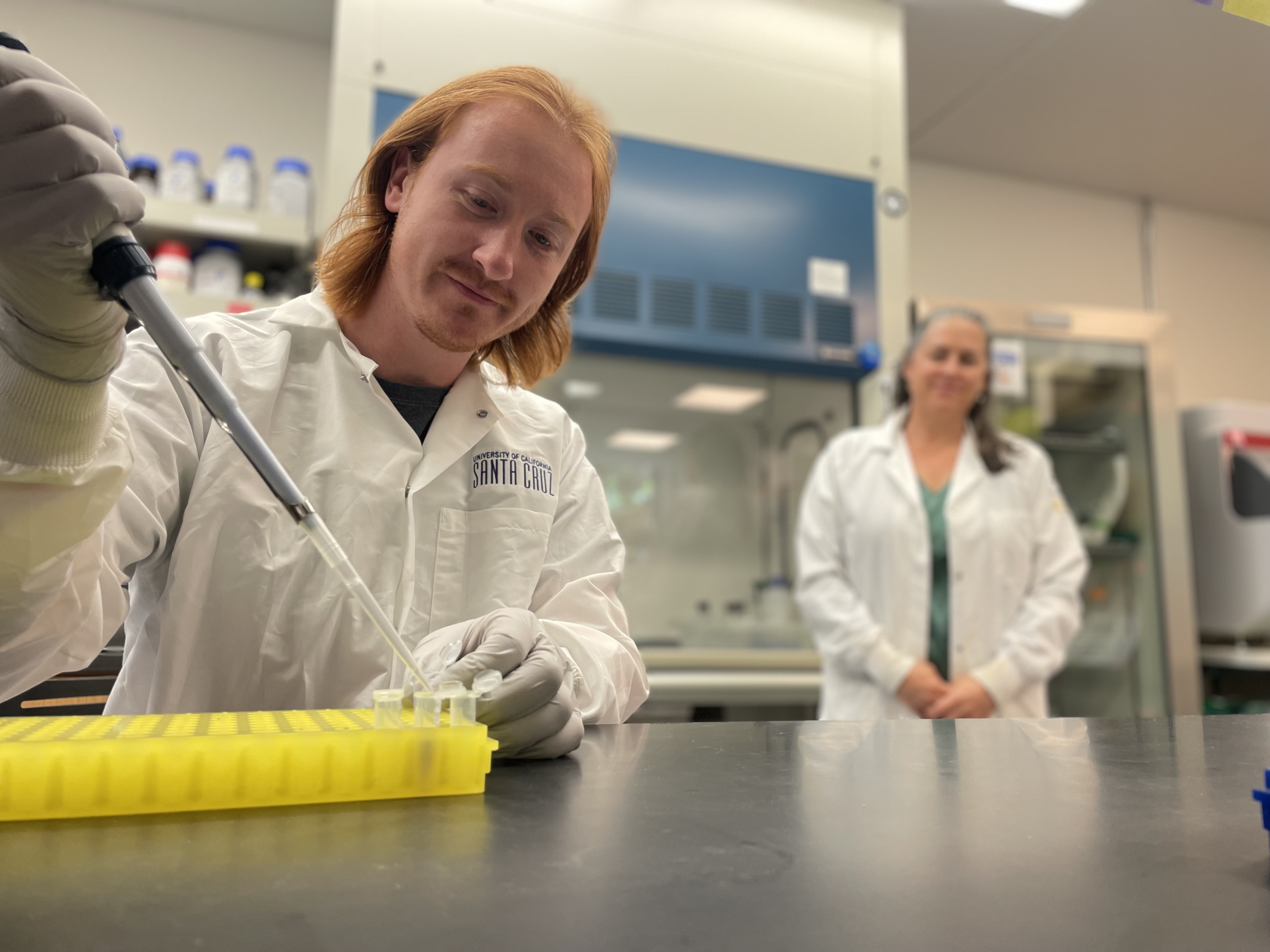 Adam Lentz pipes a pipette into a test tub at a lab table, with Professor DuBois looking on in the background.