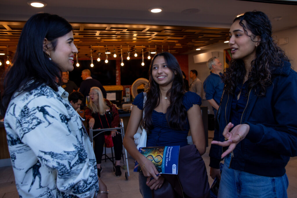 Three women talk in front of a room where others are mingling and enjoying refreshments