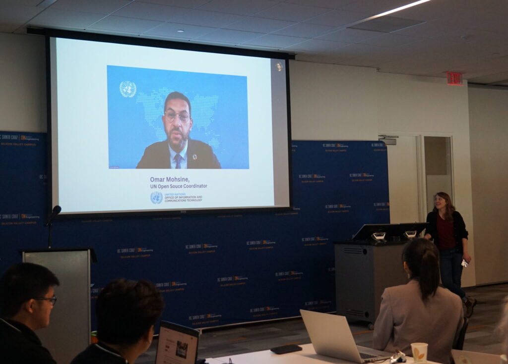 On a large screen, Omar Mohsine, UN open source coordinator, speaks to the event attendees