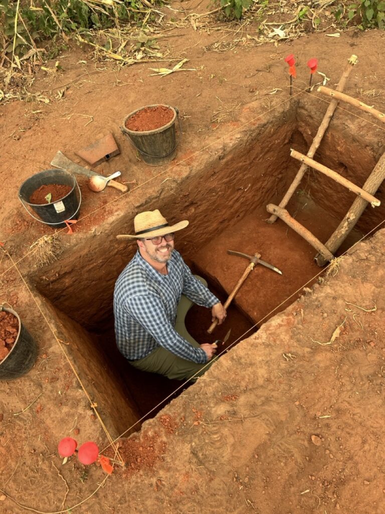 J. Cameron Monroe sits inside a red clay pit with an archaeology axe