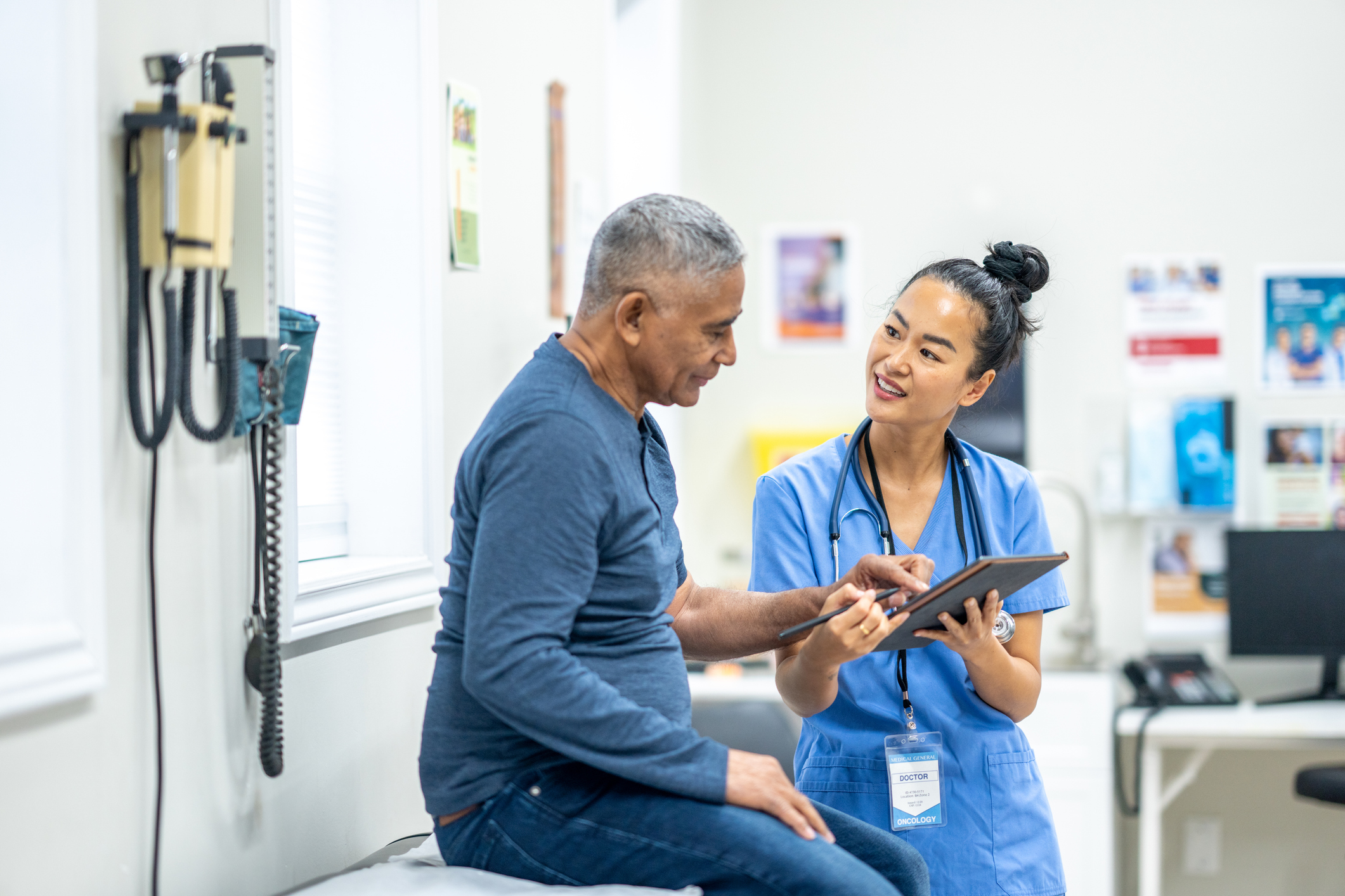 A senior gentleman of Hispanic decent, sits up on an exam table during a routine check-up at the doctors. A health care provider, who is wearing blue scrubs, is standing in front of the gentleman as she reviews some recent test results with the patient on a tablet.