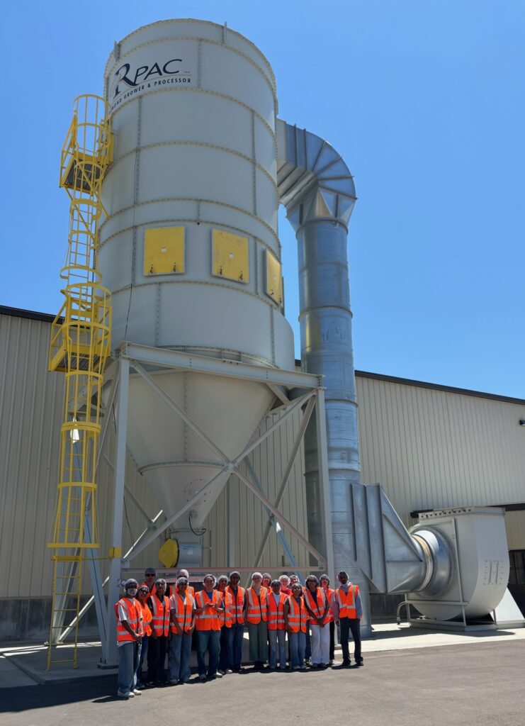 A group of people wearing orange safety vests stand in front of a large piece of machinery at the RPAC Almonds processing facility.