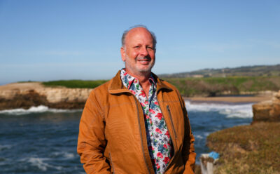 Mike Beck in a floral shirt and rust jacket by the Monterey Bay coastline.