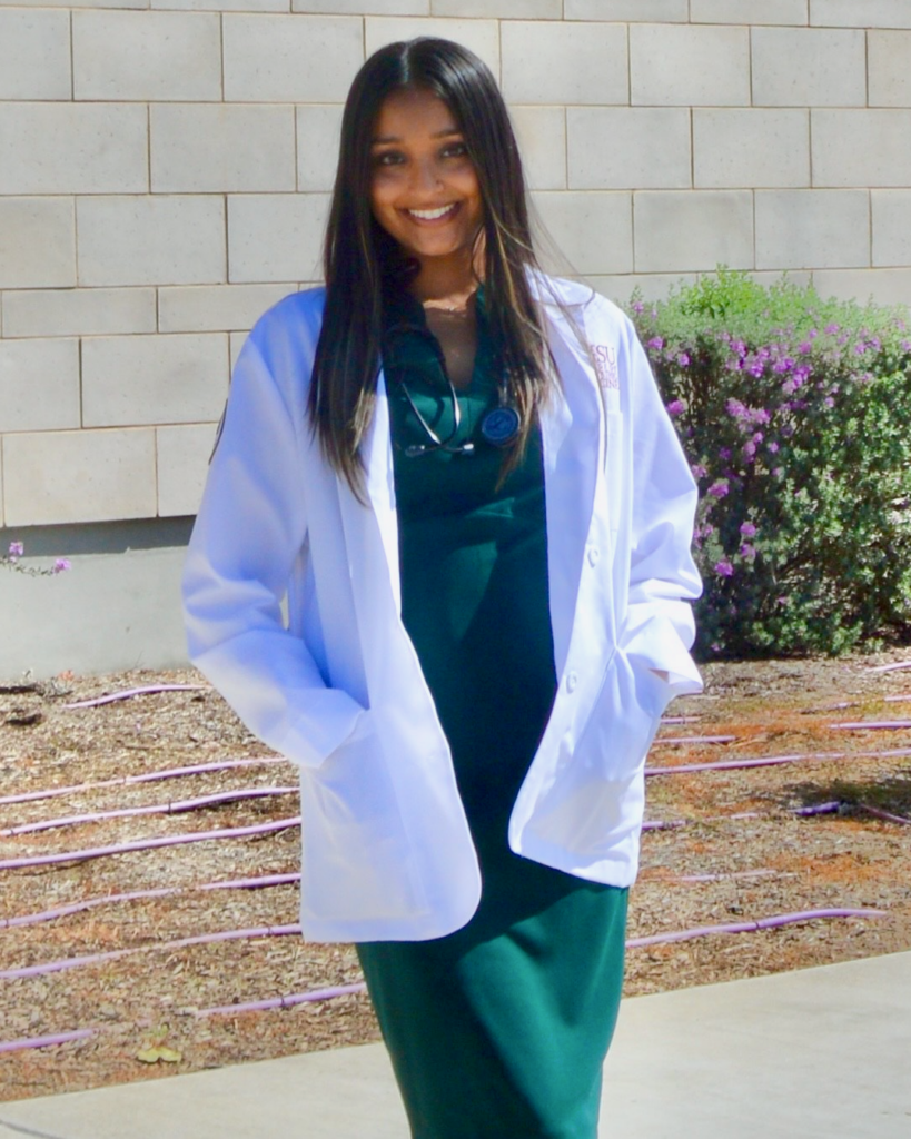 A young woman standing outside in a doctor's coat and long green dress, with a stethoscope draped around her neck. 