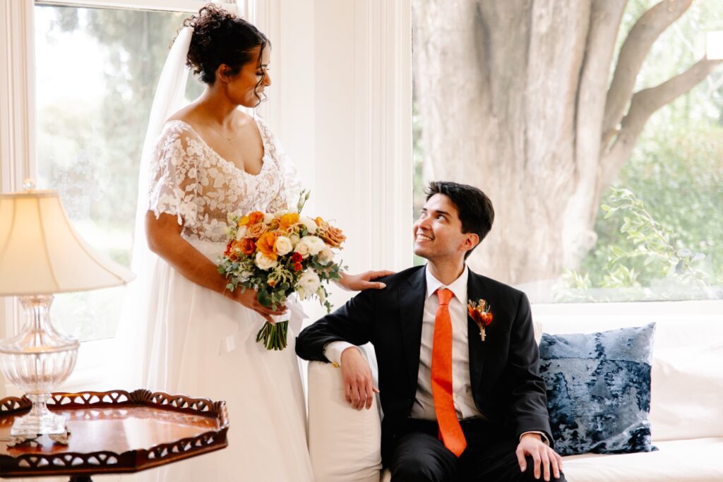 Chelsea and Brandon pose for a wedding photo. Brandon sits on a chair looking up at Chelsea, who is holding a bouquet and rests her hand on his shoulder.
