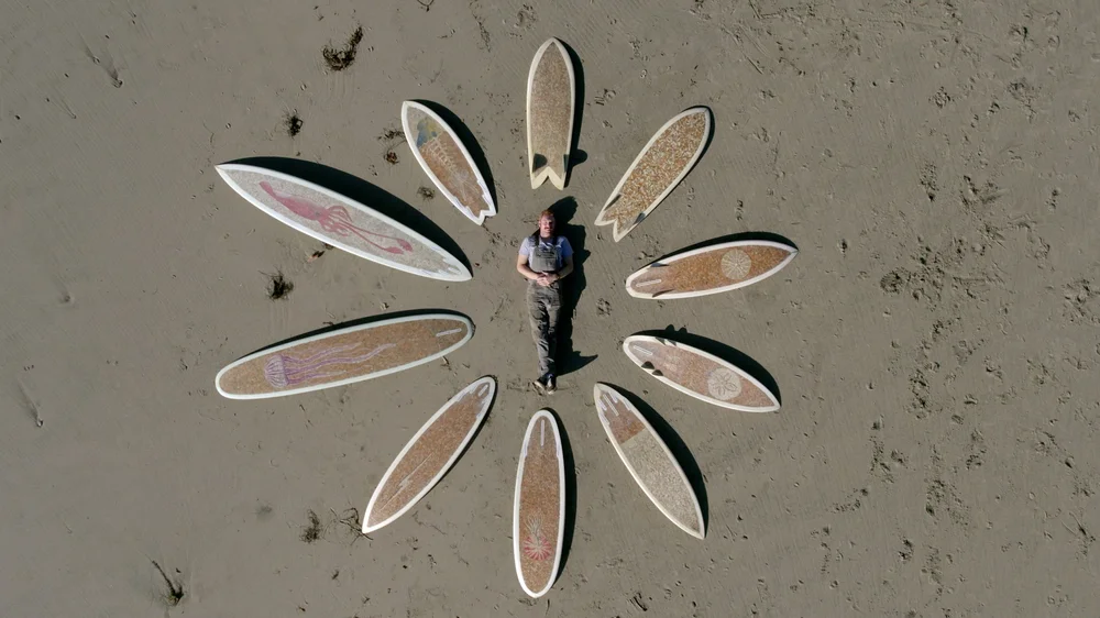 Cigarette surfboard creator Taylor Lane lays in the sand surrounded by 10 cigarette surfboards.