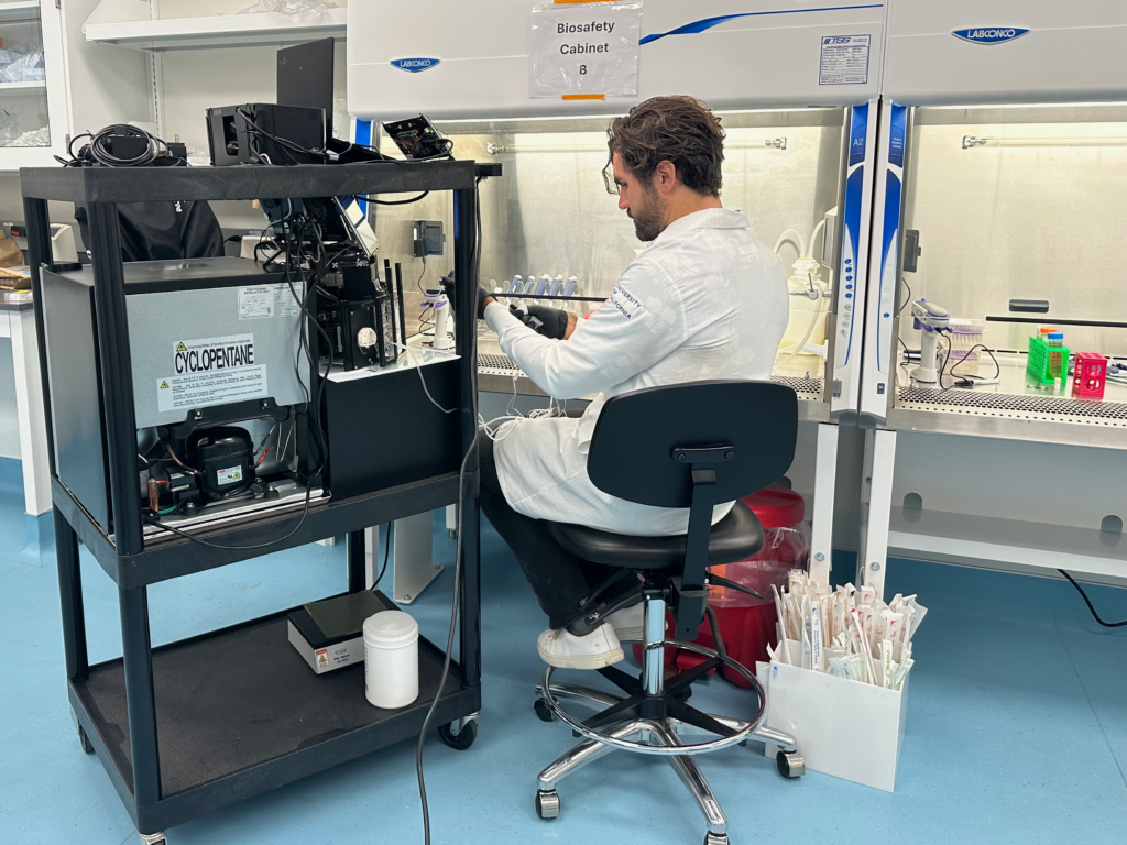 Seiler in a lab coat with his back to the camera, sits next to a portable metal shelf with multiple pieces of scientific equipment on it.