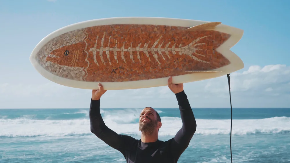 A surfer holds a cigarette surfboard above his head on the beach.