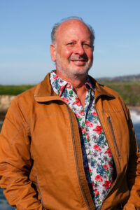 Mike Beck in a floral patterned shirt and rust jacket outside by the Monterey Bay coastline.