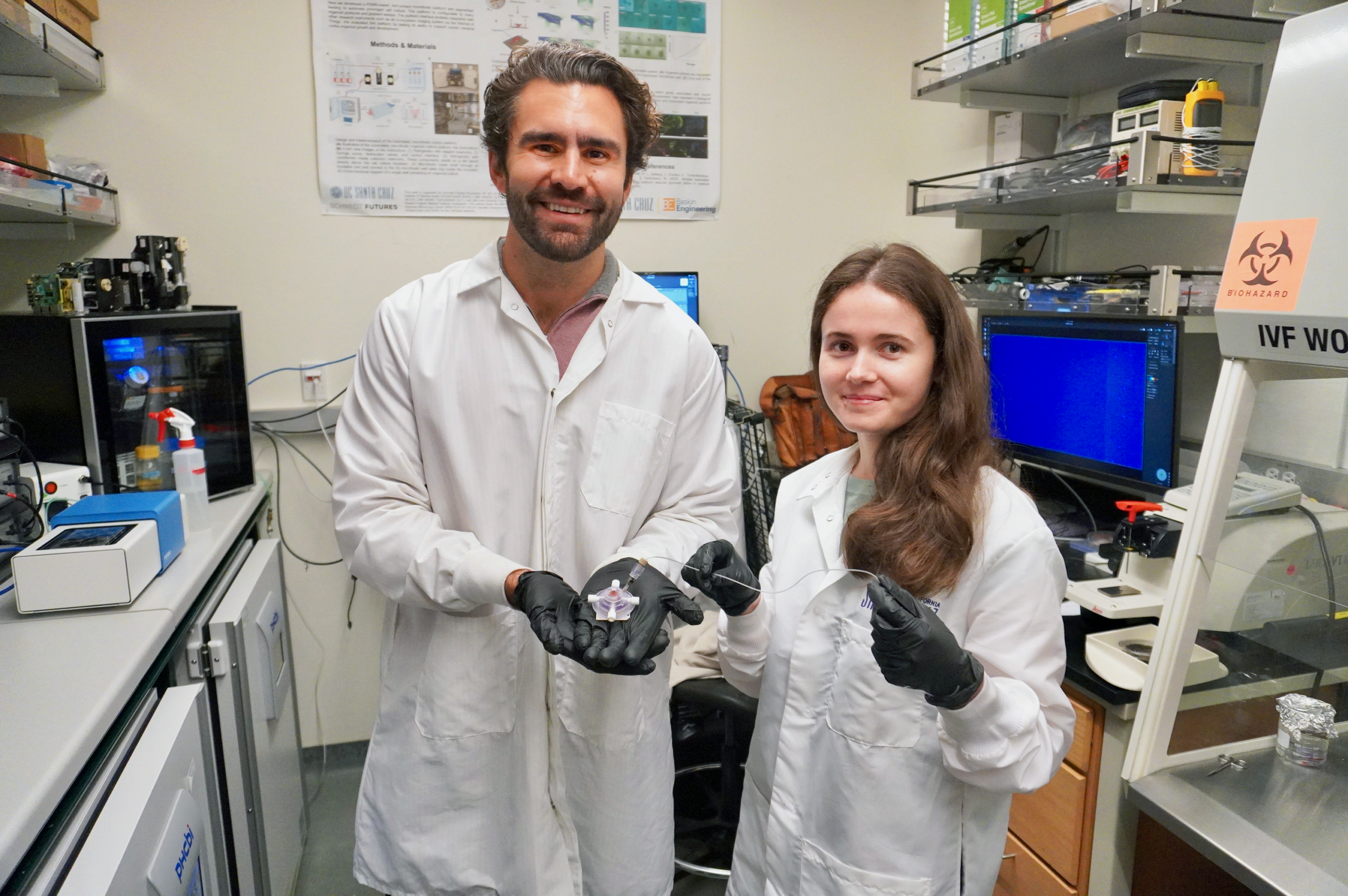 A male and female scientist in lab coats standing in a lab. The man is holding a small circular device in open hands.