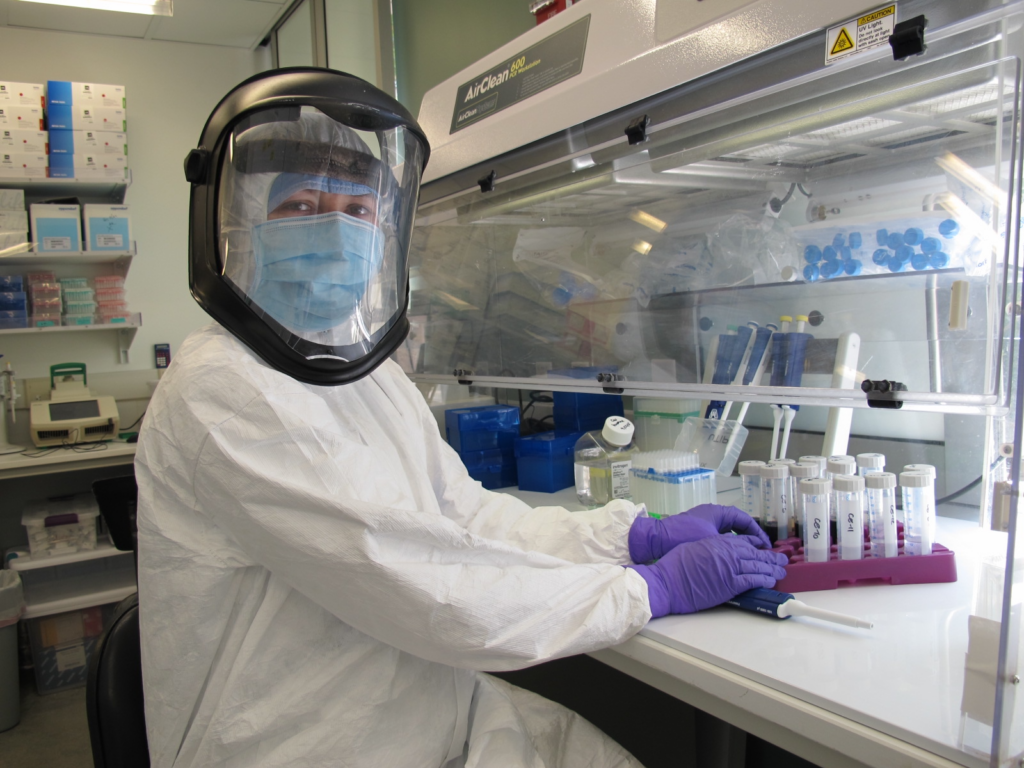 A woman in a full-body cleanroom suit and gloves holds samples under a hood.