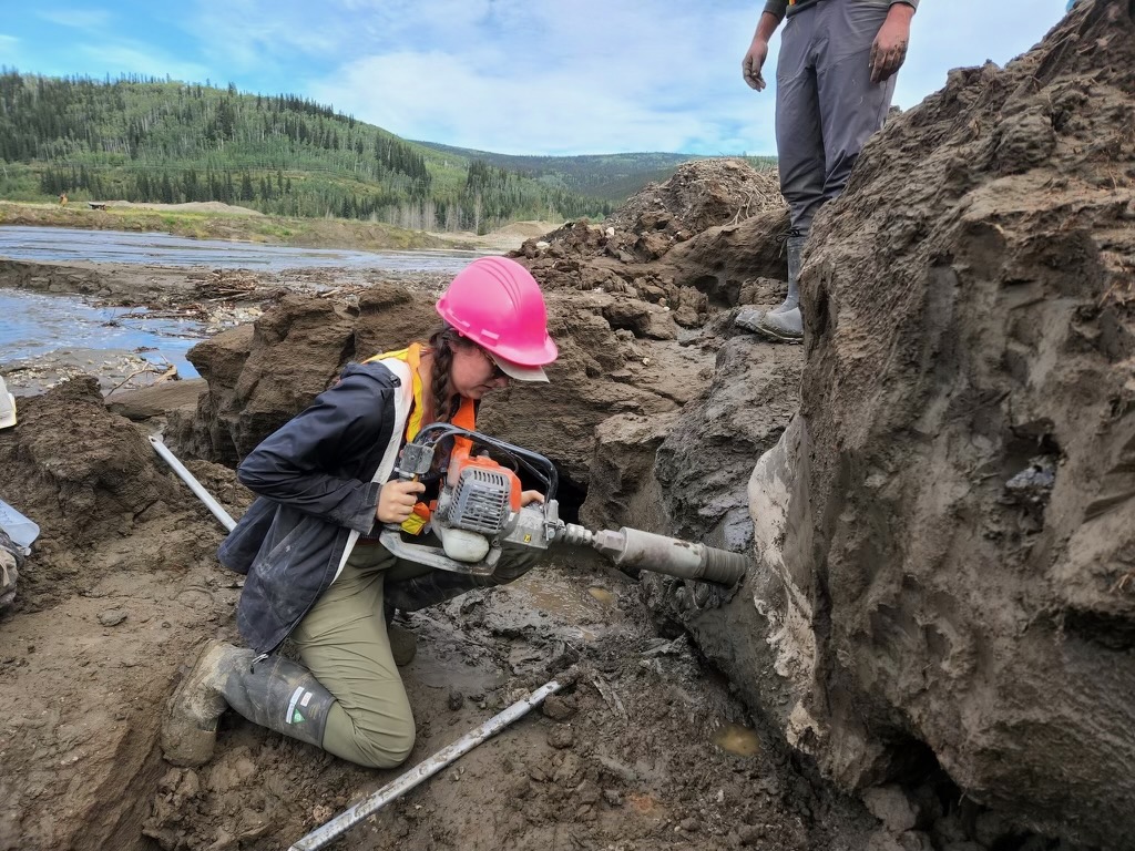 A young woman in a hardhat holds a large drill to the side of a mound of compressed sediment as someone stands above.
