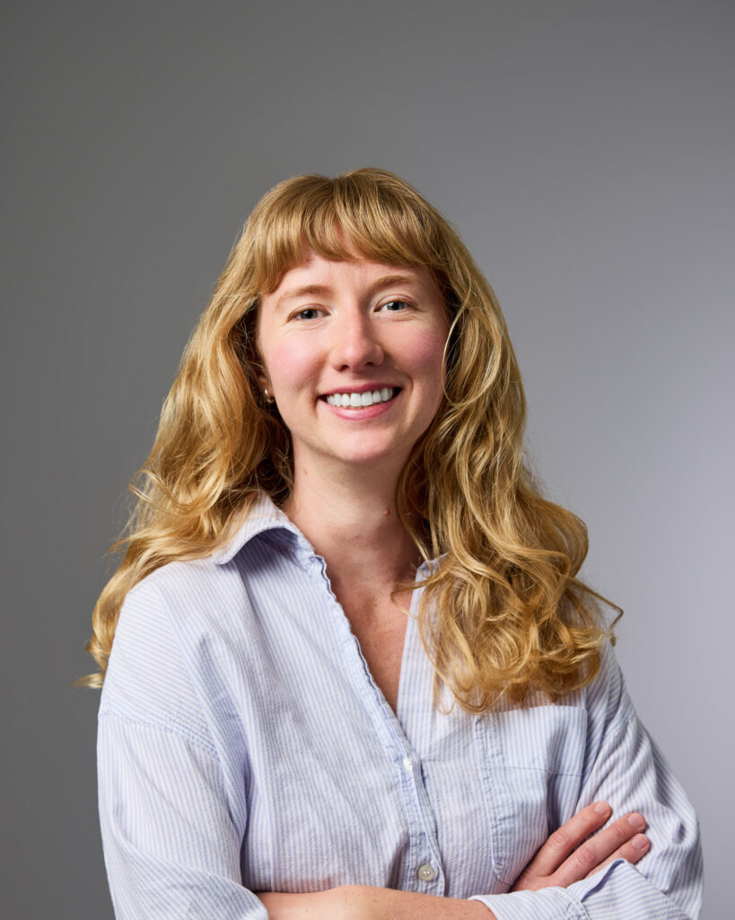 Woman with long blond hair and bangs and a big smile, crossing her arms in front of her. 