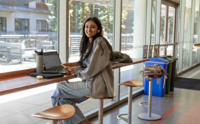 Student smiles while sitting at a long table with a laptop in front of her.