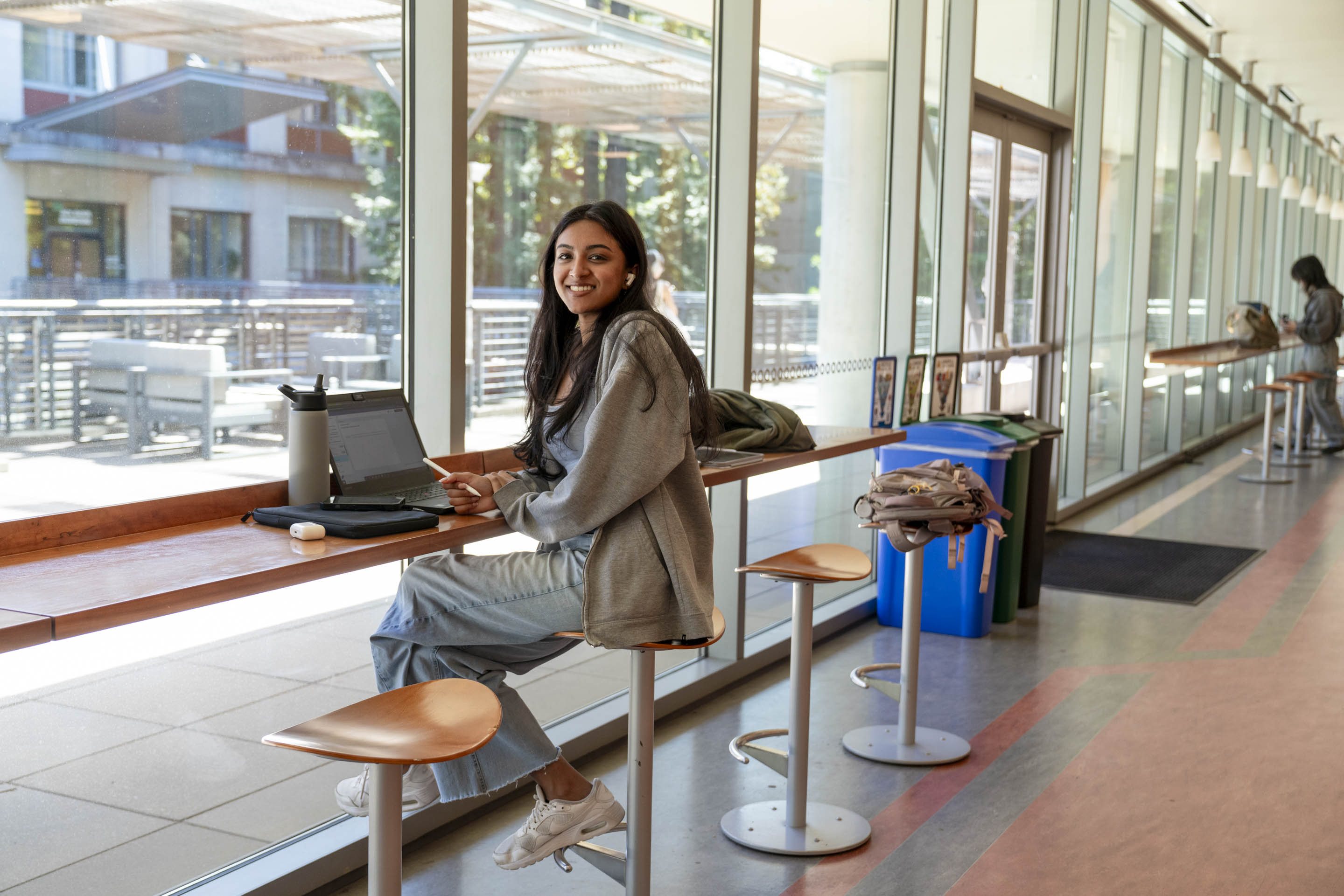 Student smiles while sitting at a long table with a laptop in front of her.