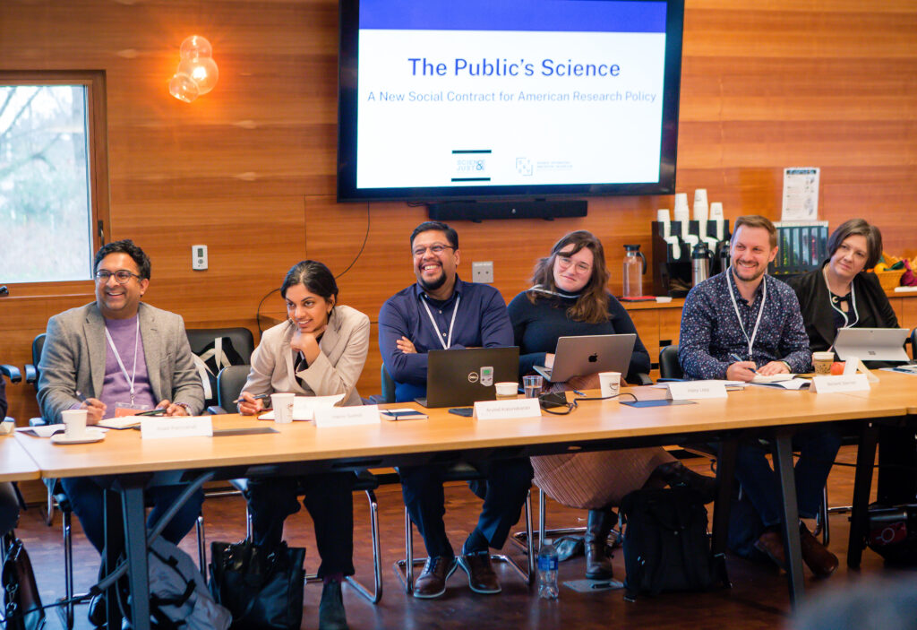 Five people smile while sitting at a long table. A screen behind reads "The Public's Science"
