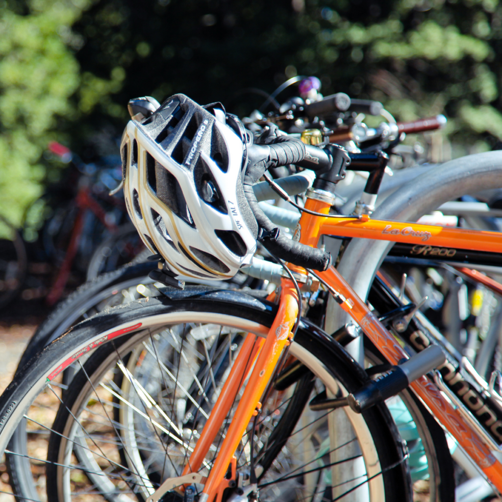 Orange bicycle parked in a rack with a white helmet resting on the handlebars, surrounded by other bikes in a shaded outdoor area.