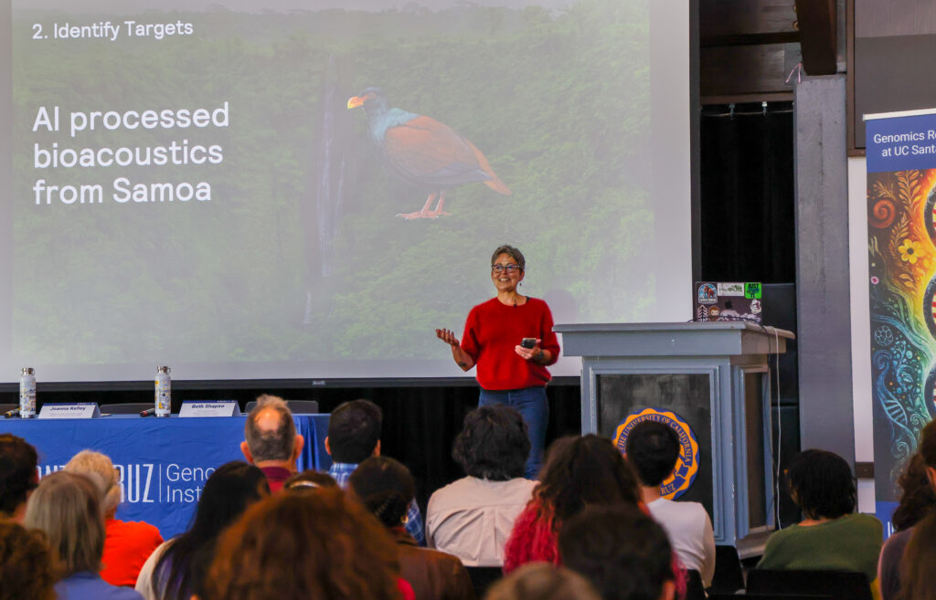 Beth Shapiro stands next to a podium in front of a crowd of people, with a slide of an exotic bird behind her