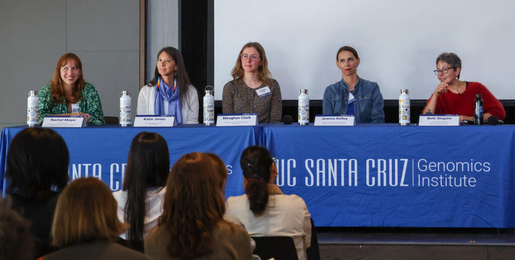 Five women sit facing the audience behind a table with a bright blue tablecloth labeled "UC Santa Cruz Genomics Institute"