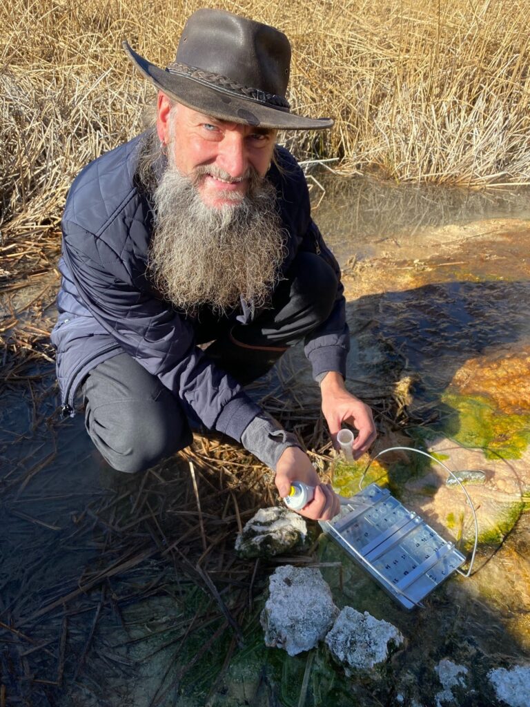 Damer squats near a shallow stream as he pipettes liquid into a vial
